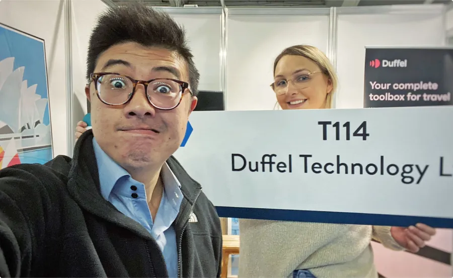 Man taking selfie of himself and woman in a conference booth holding up a Duffel sign.