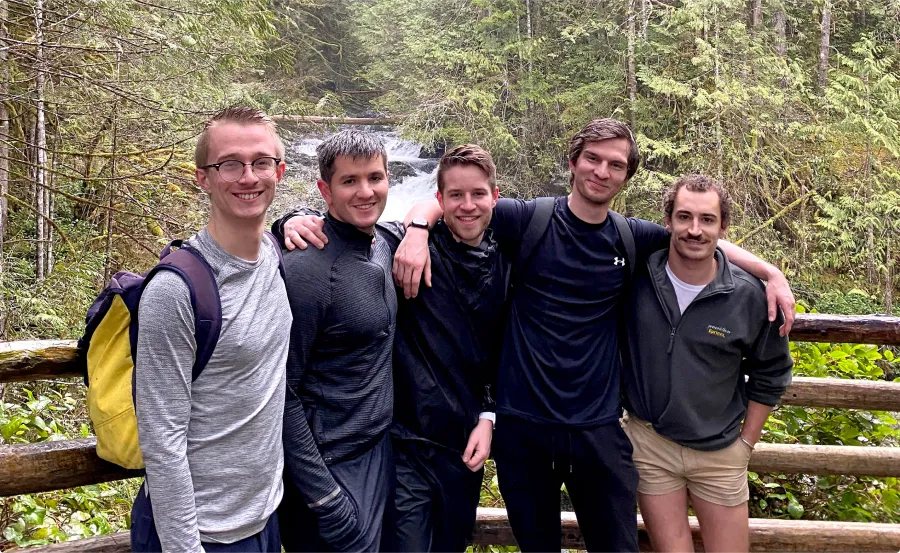 Five men hugging and posing for picture on a bridge over a river.