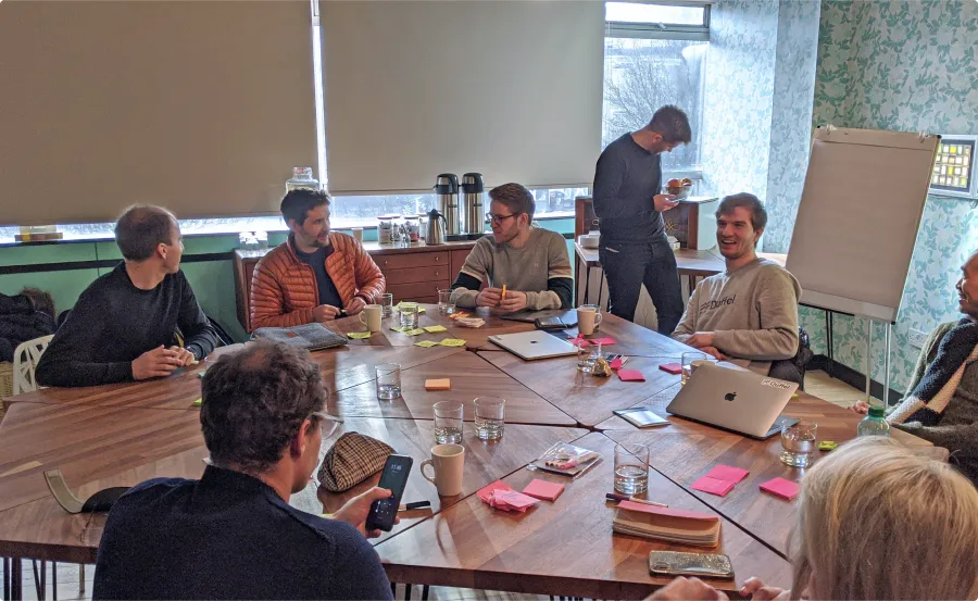 Seven people sitting around a table talking, writing and smiling with a man standing next to a white paper board next to them.